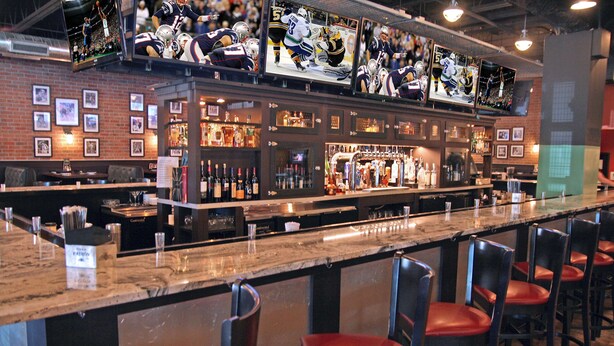 Zona de bar con una barra larga, varios televisores mostrando deportes y diferentes opciones de asientos.
