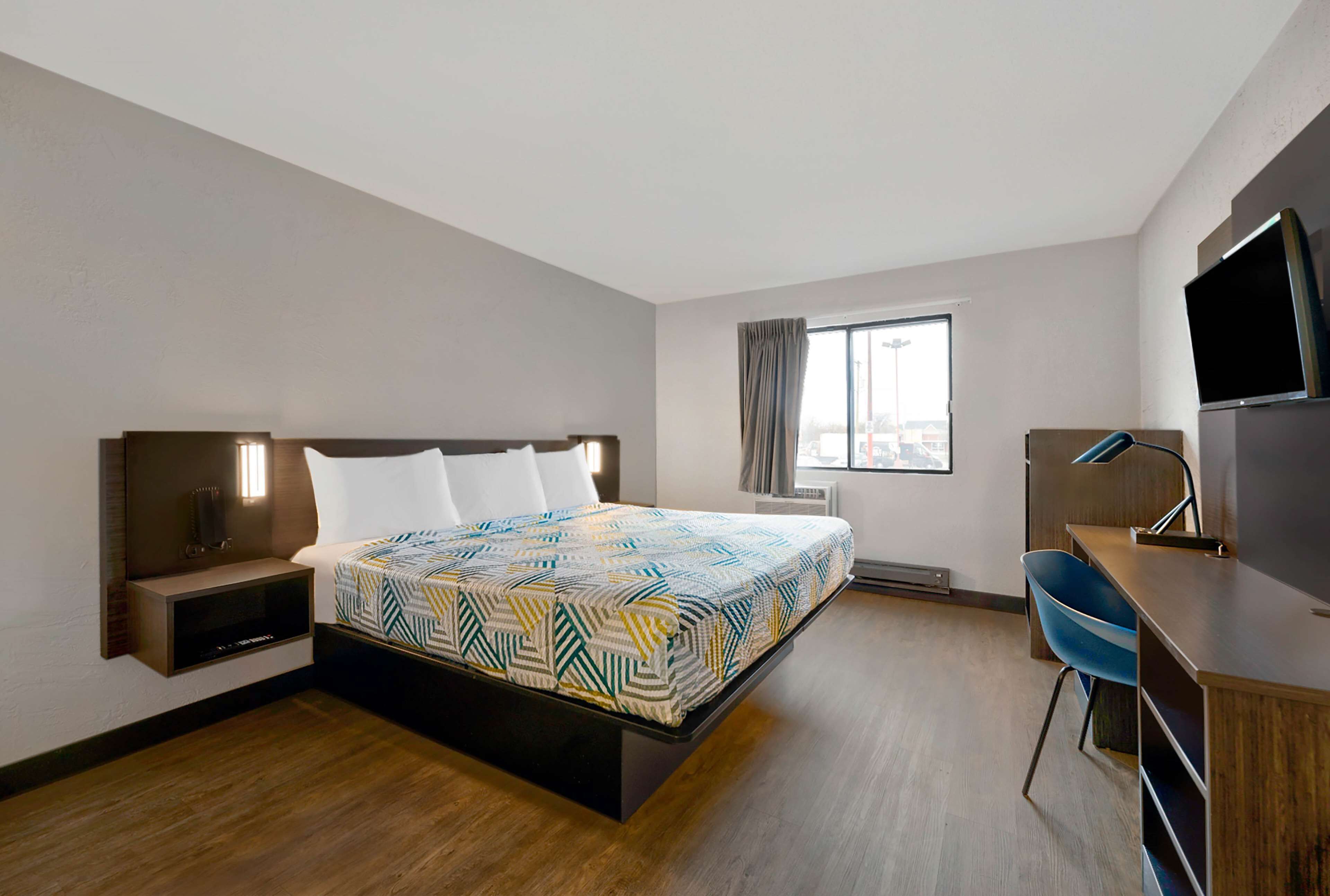 A hotel room with a bed, bedside table, TV, desk, chair, and a window with curtains.