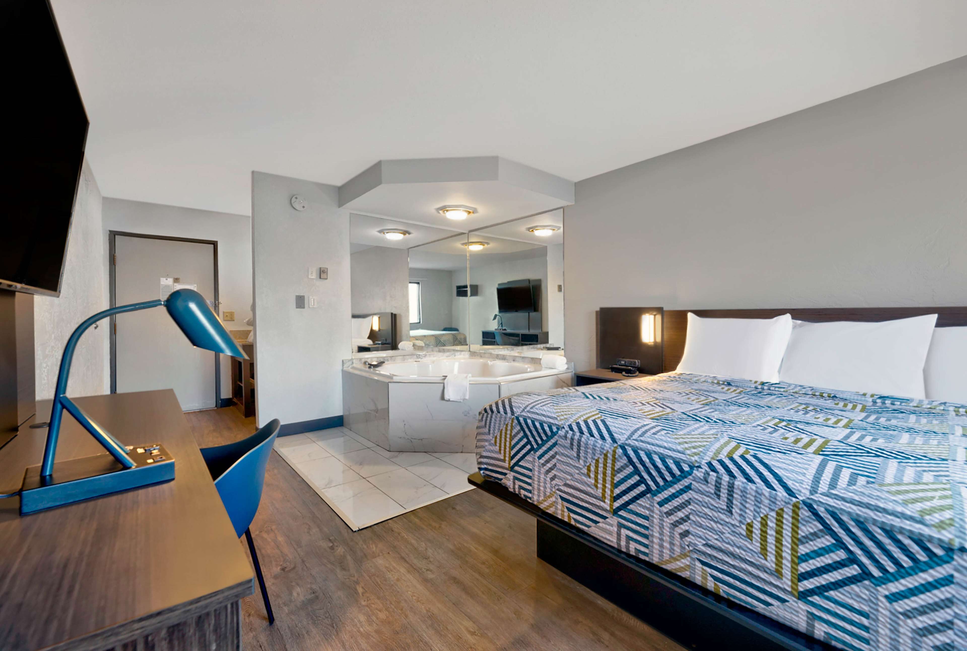 A hotel room with a bed, a desk, a chair, a desk lamp, a large mirror, and a bathroom with a bathtub and sink.