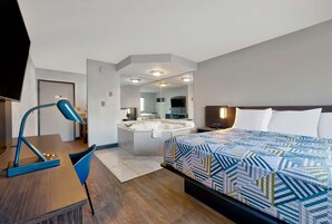 A hotel room with a bed, a desk, a chair, a desk lamp, a large mirror, and a bathroom with a bathtub and sink.