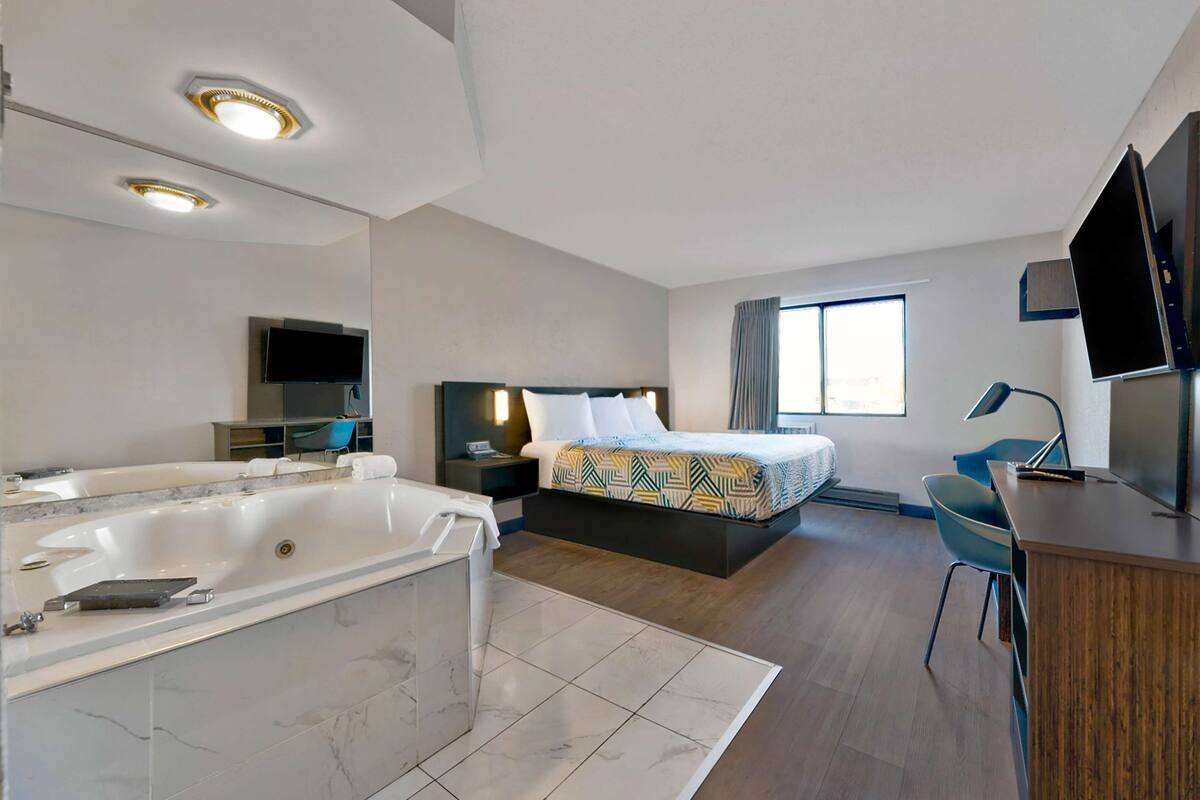 A modern hotel room with a large bathtub, a bed with a patterned bedspread, a desk with a computer, and a TV mounted on the wall.