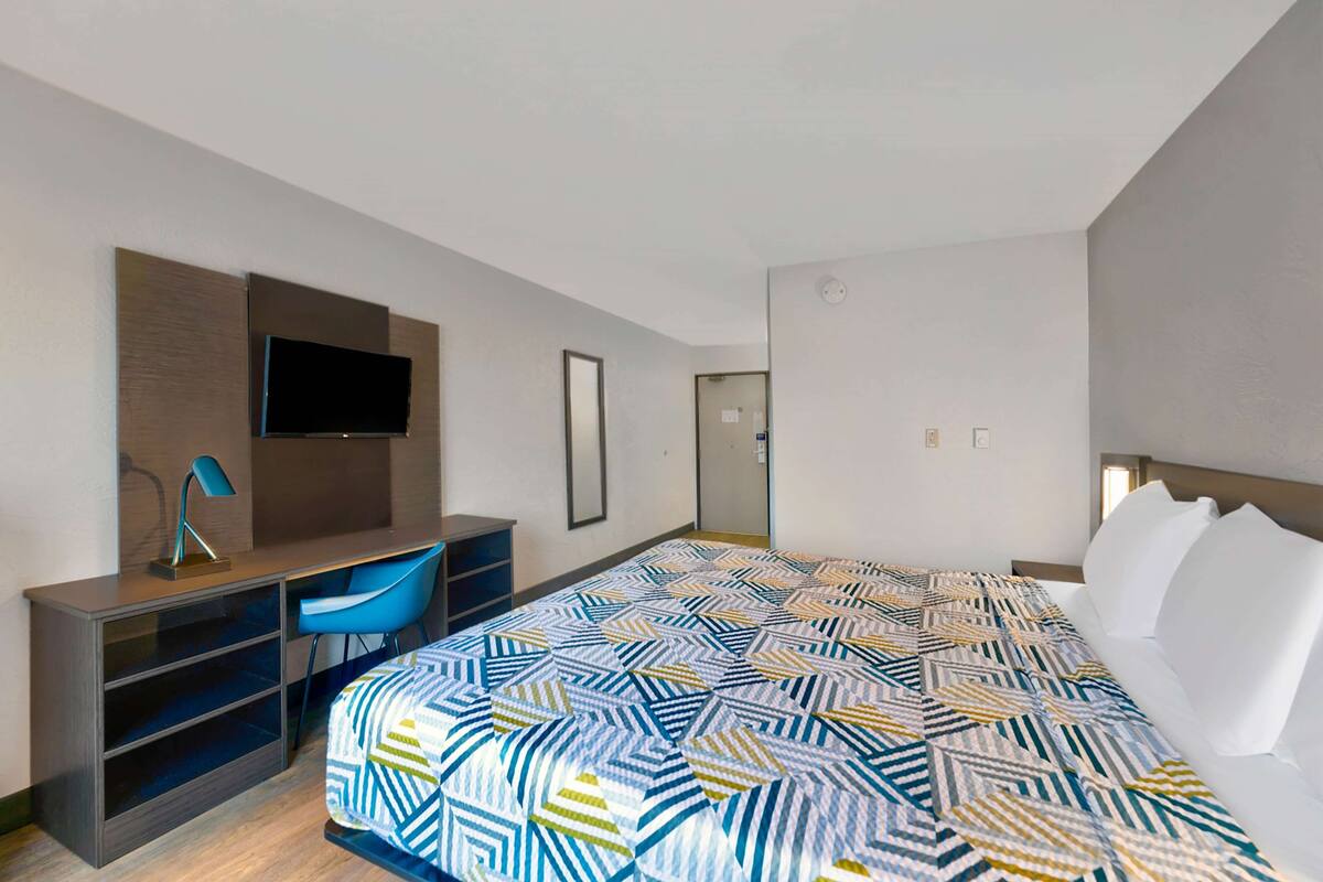 A modern hotel room with a large bed, a desk with a chair, a flat-screen TV mounted on the wall, and a door leading to another room.