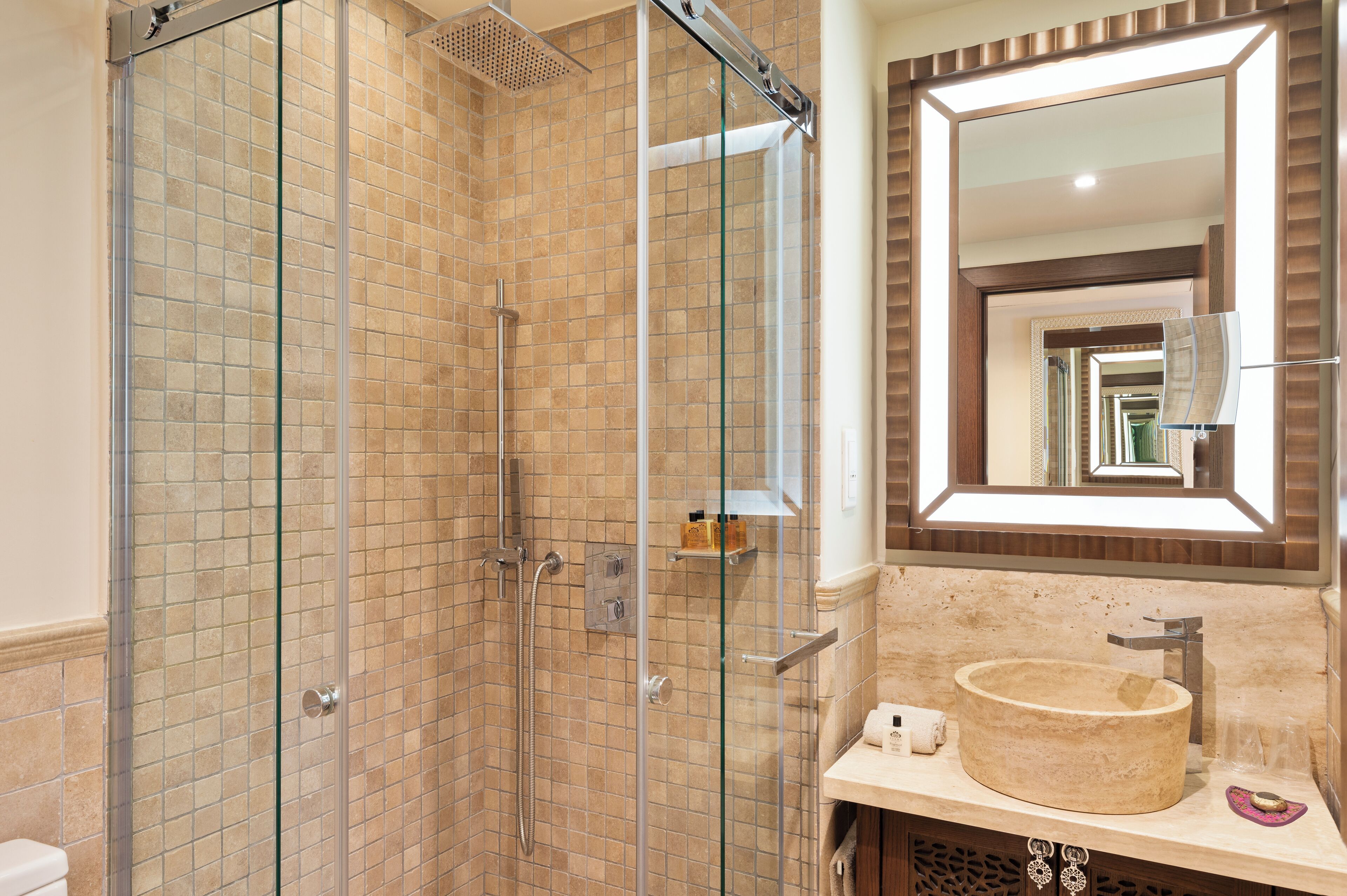 A modern bathroom with a glass-enclosed shower, a beige tiled wall, a large mirror, and a round sink.
