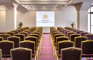 Un salón de conferencias con filas de sillas moradas y una pantalla grande que muestra el nombre del hotel.