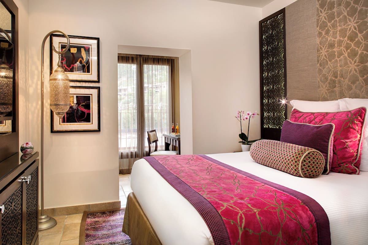A hotel room with a large bed, a desk, and a chair. There are two framed pictures on the wall, a window with curtains, and a decorative wall panel.