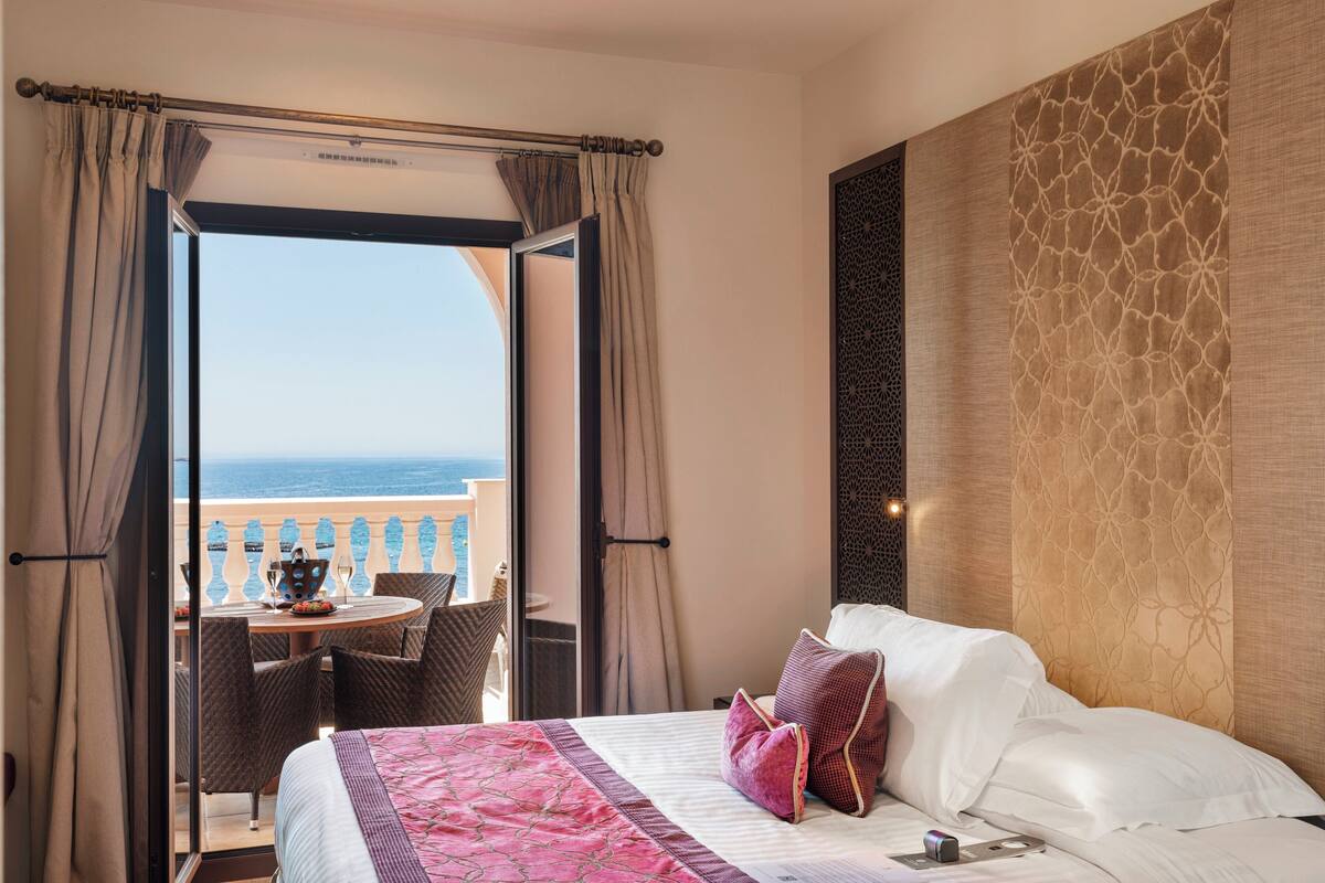 A hotel room with a balcony, a bed with white linens, a pink and white patterned bedspread, and a view of the sea.