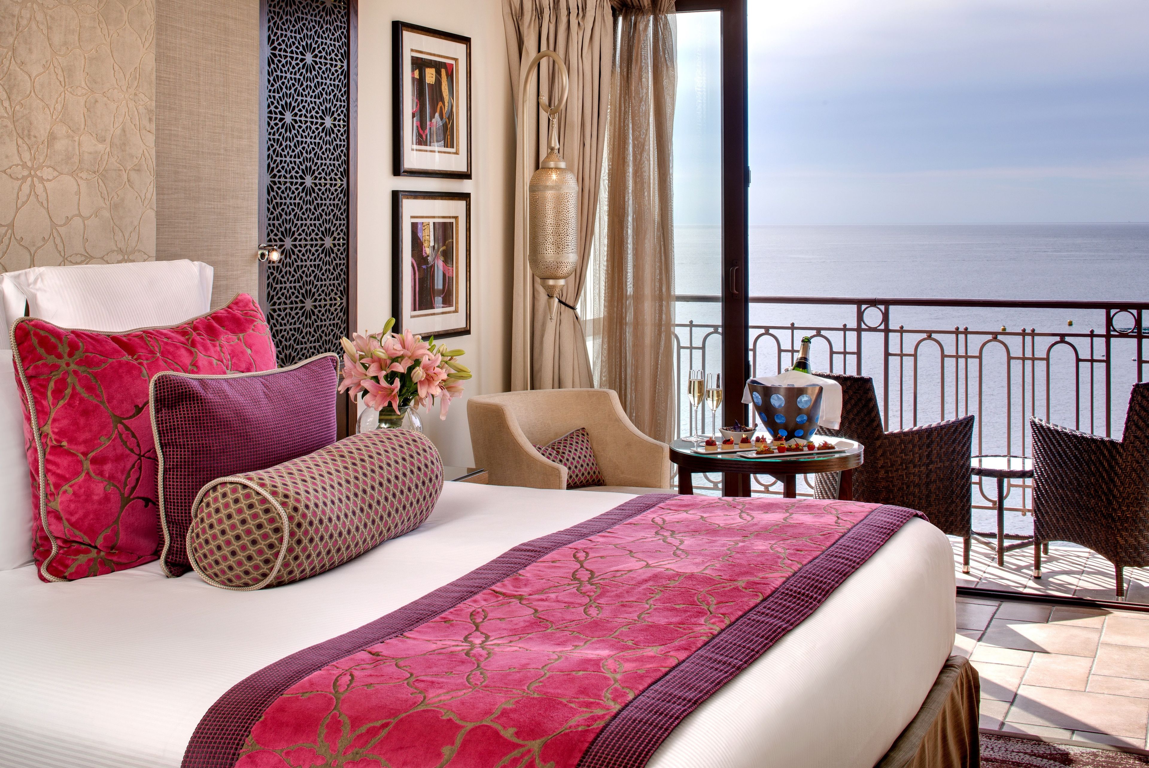 Een hotelkamer met een bed, uitzicht op zee, een balkon met stoelen en een tafel met een bloevas.