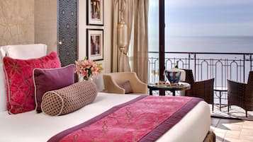 Een hotelkamer met een bed, uitzicht op zee, een balkon met stoelen en een tafel met een bloevas.