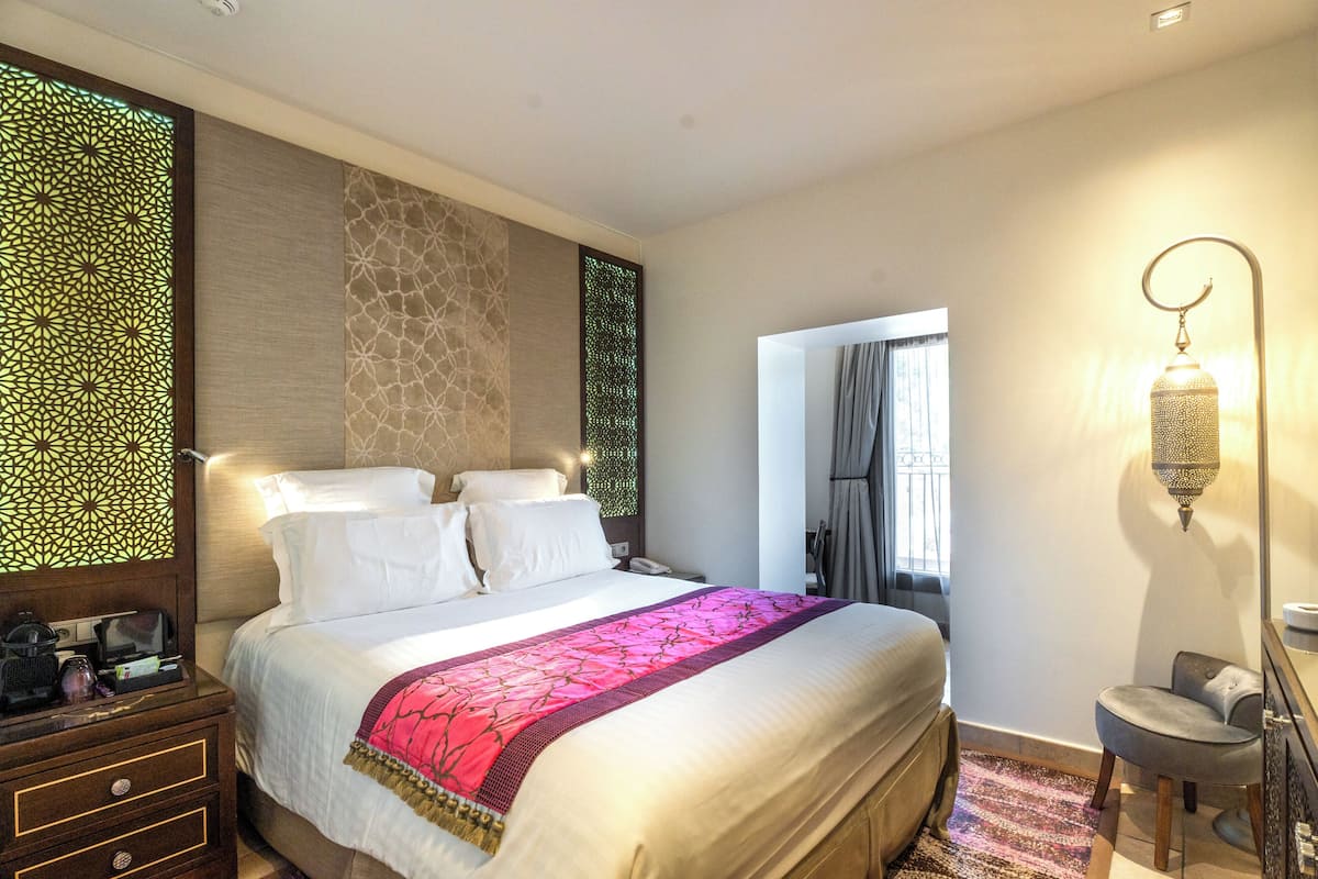 Habitación de hotel luminosa con una cama grande, una mesita de noche con lámpara y vistas al exterior a través de una ventana.