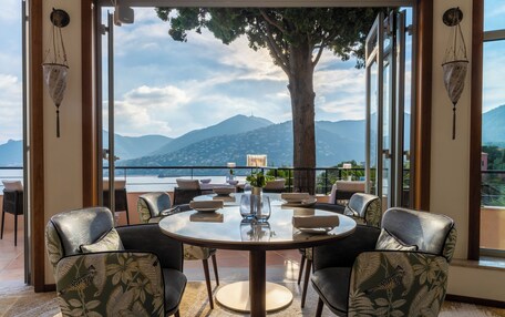 A round table with chairs, a vase with flowers, and a view of mountains and a lake.