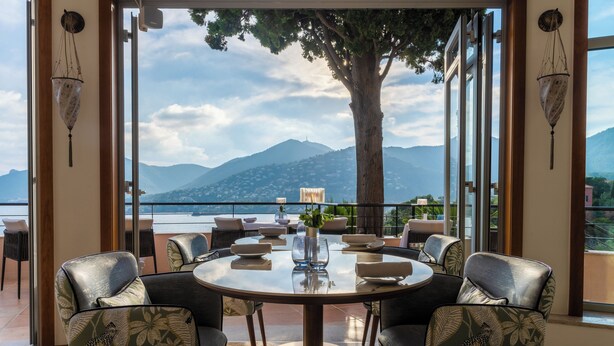 A round table with chairs, a vase with flowers, and a view of mountains and a lake. - Tiara Miramar Beach Resort (Theoule-sur-Mer)