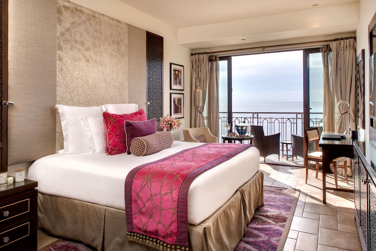A spacious bedroom with a large bed, a balcony with a view of the sea, and a dining area with chairs.