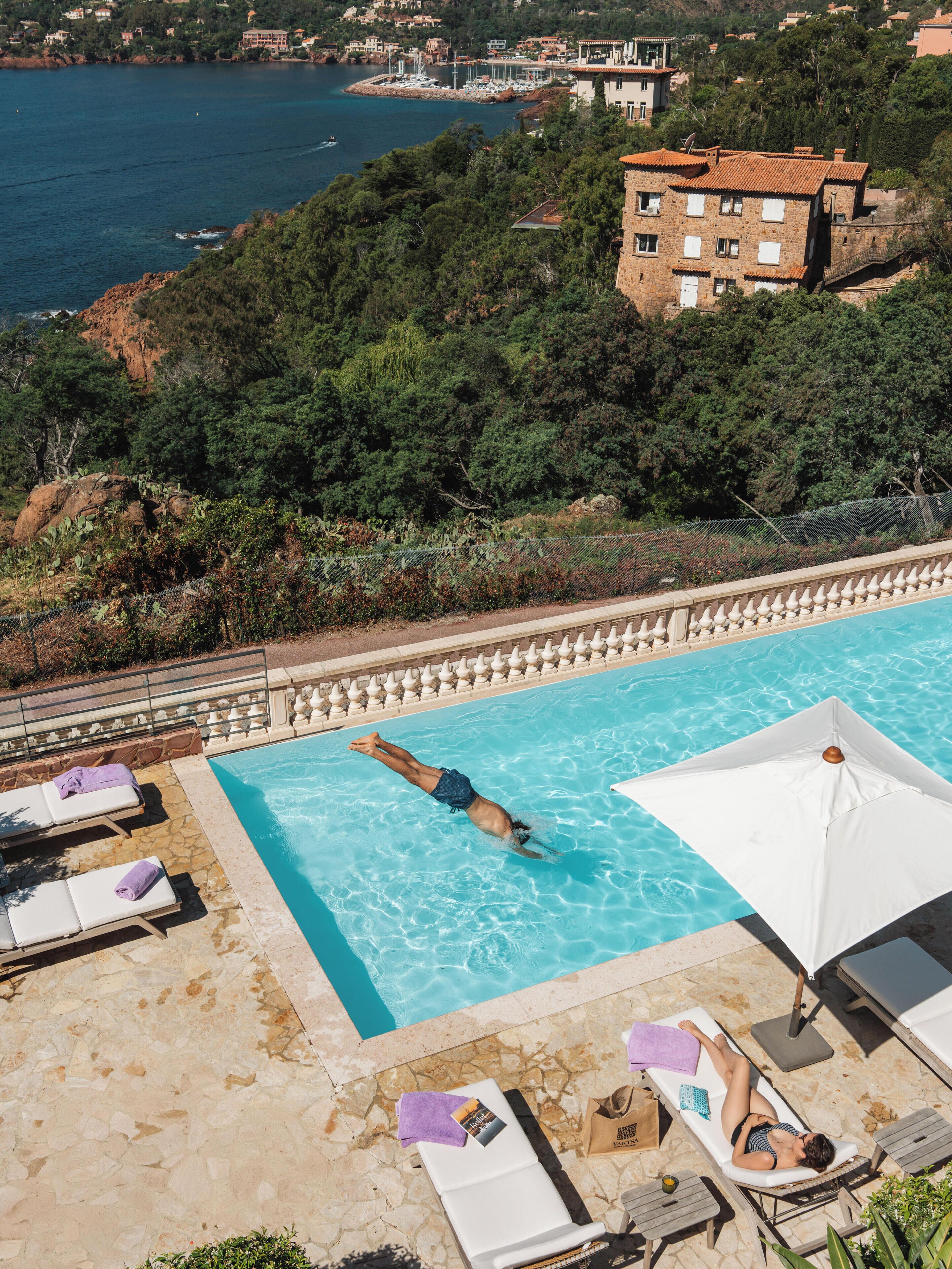 Una piscina con tumbonas y sombrillas, rodeada por una terraza de piedra y vistas a una zona costera con edificios y árboles.