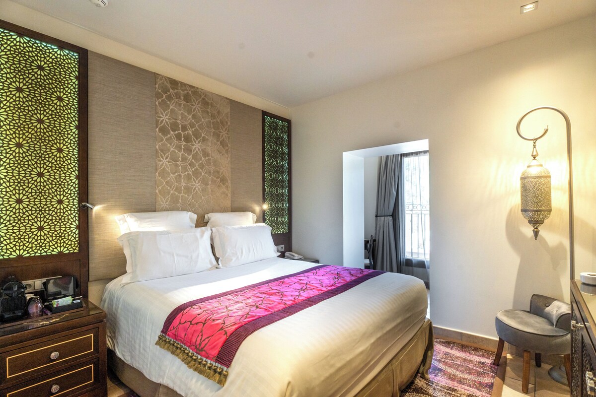 A well-lit hotel room with a large bed, a bedside table with a lamp, and a view of the outside through a window.