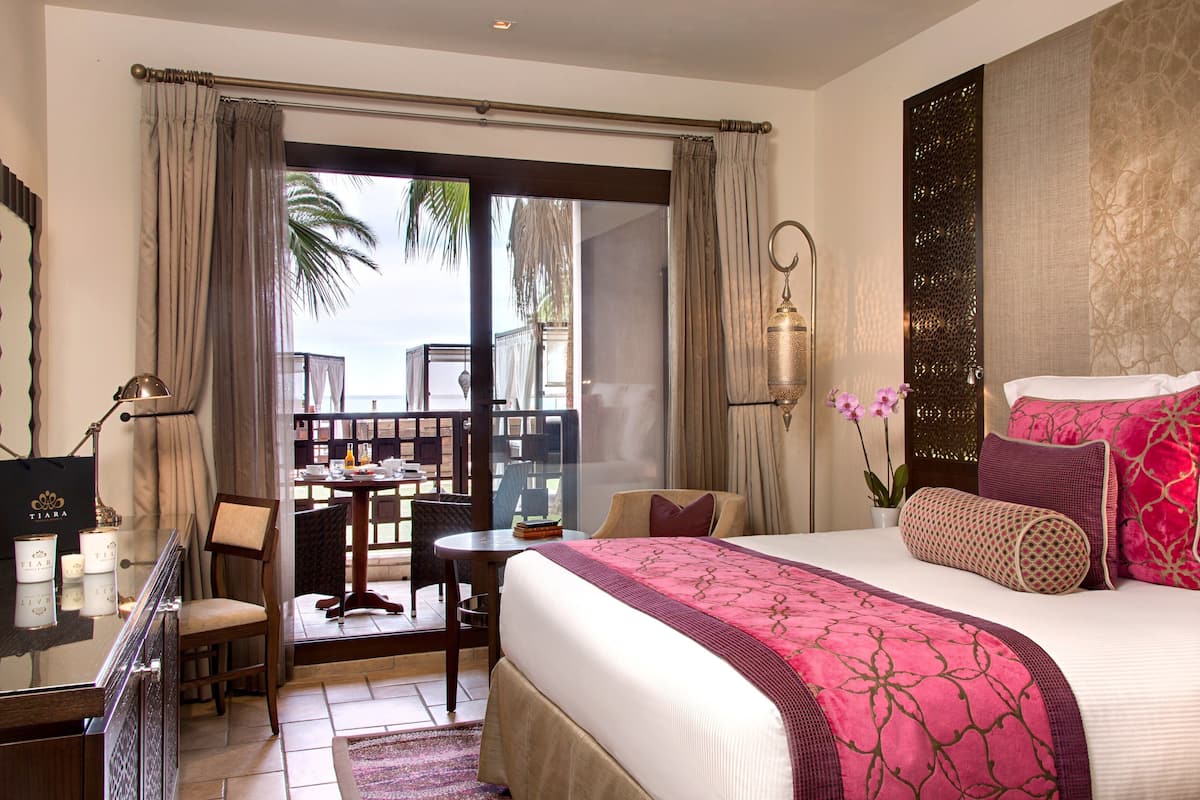 Habitación de hotel con una cama grande, un escritorio y una silla. Tiene un balcón con vistas a palmeras.