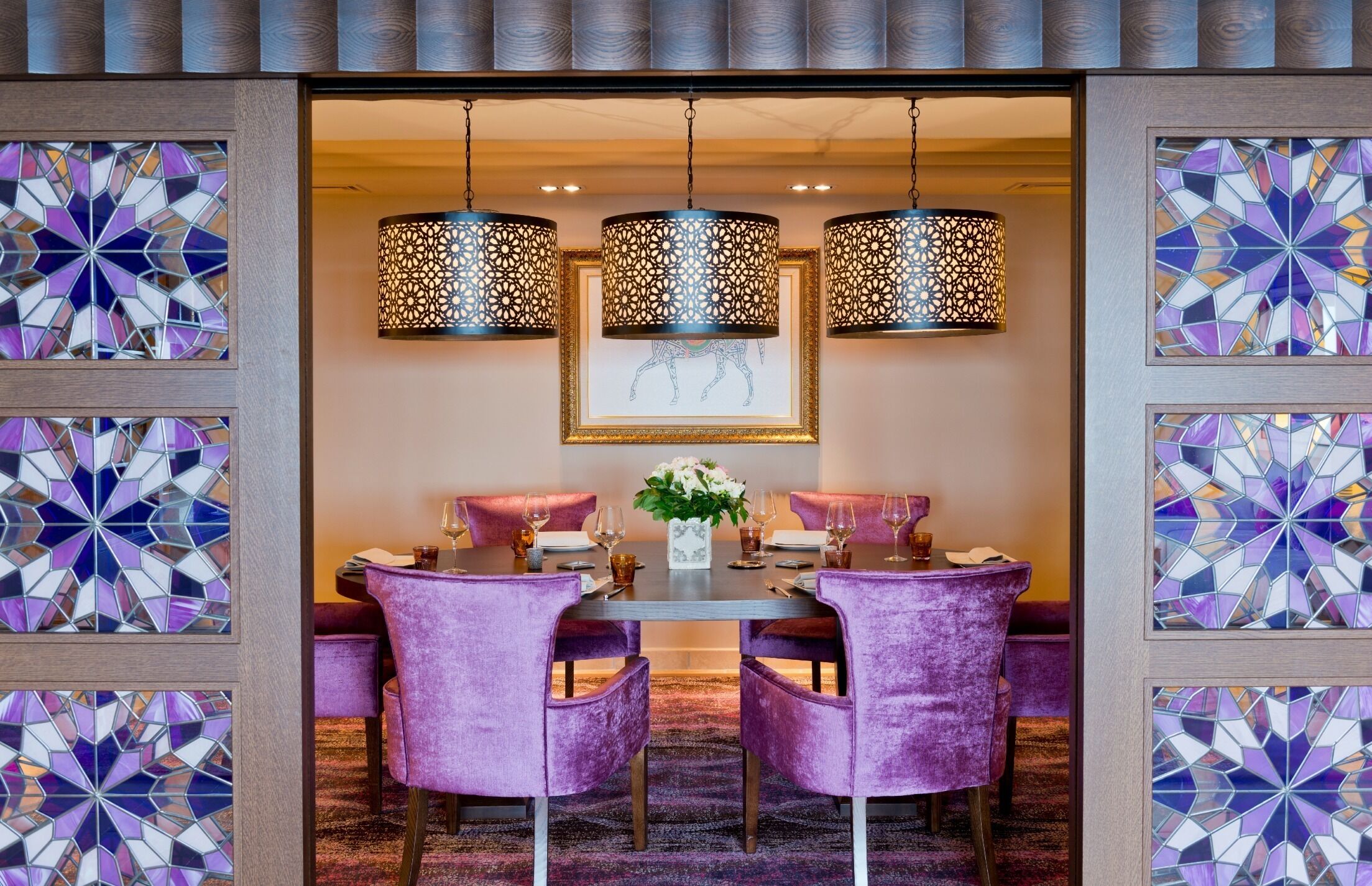 Un comedor con una mesa redonda, sillas tapizadas en color morado y lámparas colgantes con diseños elaborados.