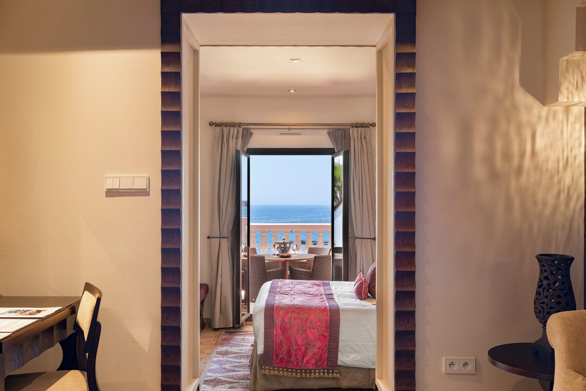 A hotel room with a bed, a desk, and a view of the sea through a balcony.