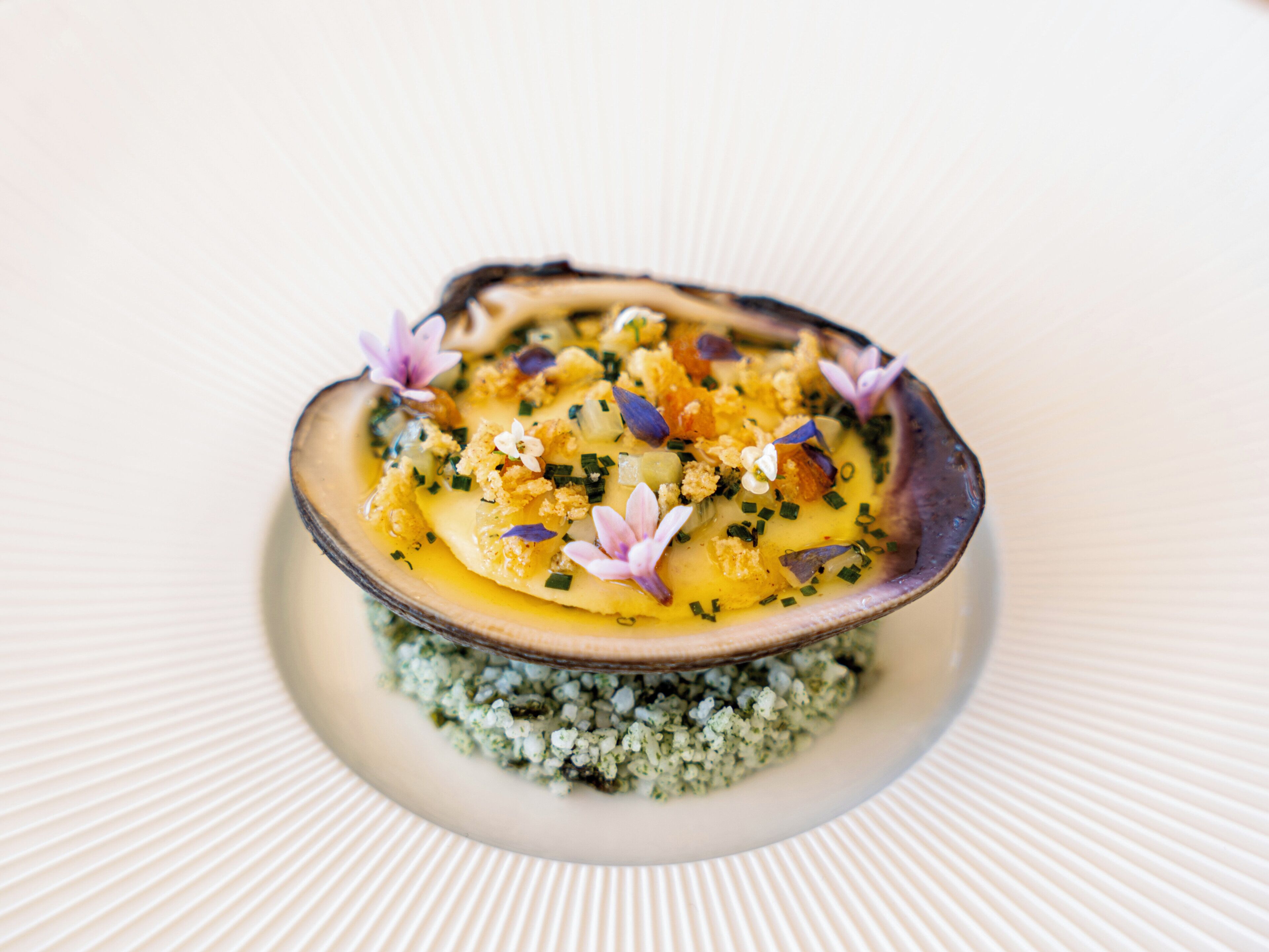 A dish with a creamy base, garnished with purple flowers and green herbs, served on a textured white plate.