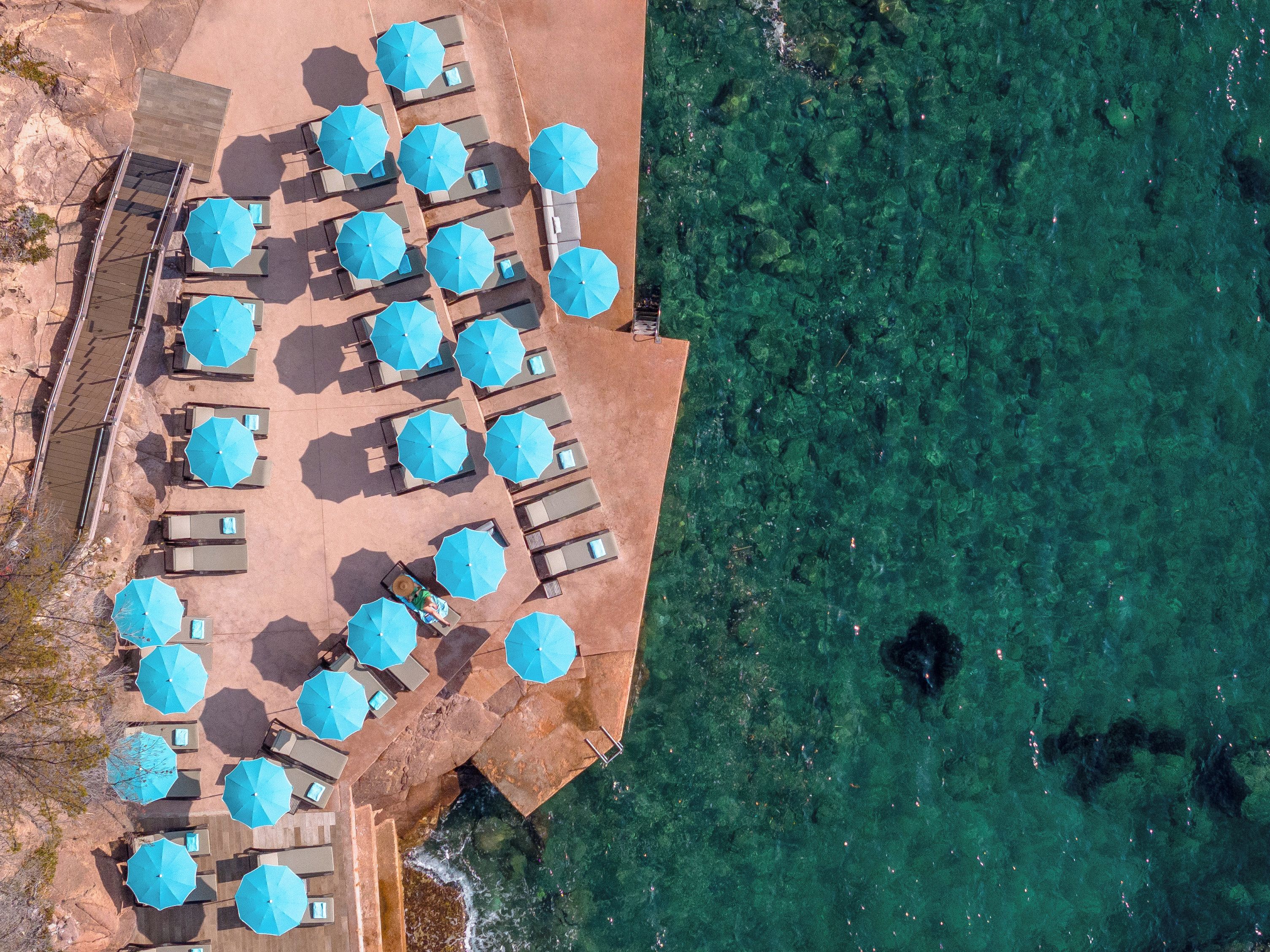 An aerial view of a beachside resort with multiple sun loungers and umbrellas arranged in rows, overlooking clear turquoise waters.