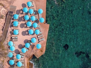 An aerial view of a beachside resort with multiple sun loungers and umbrellas arranged in rows, overlooking clear turquoise waters. - Tiara Miramar Beach Resort (Theoule-sur-Mer)