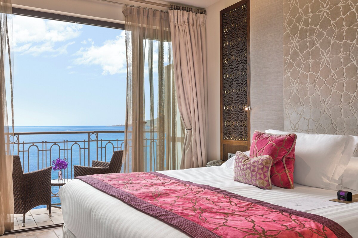 A hotel room with a large bed, a view of the sea, and a balcony with wicker chairs.