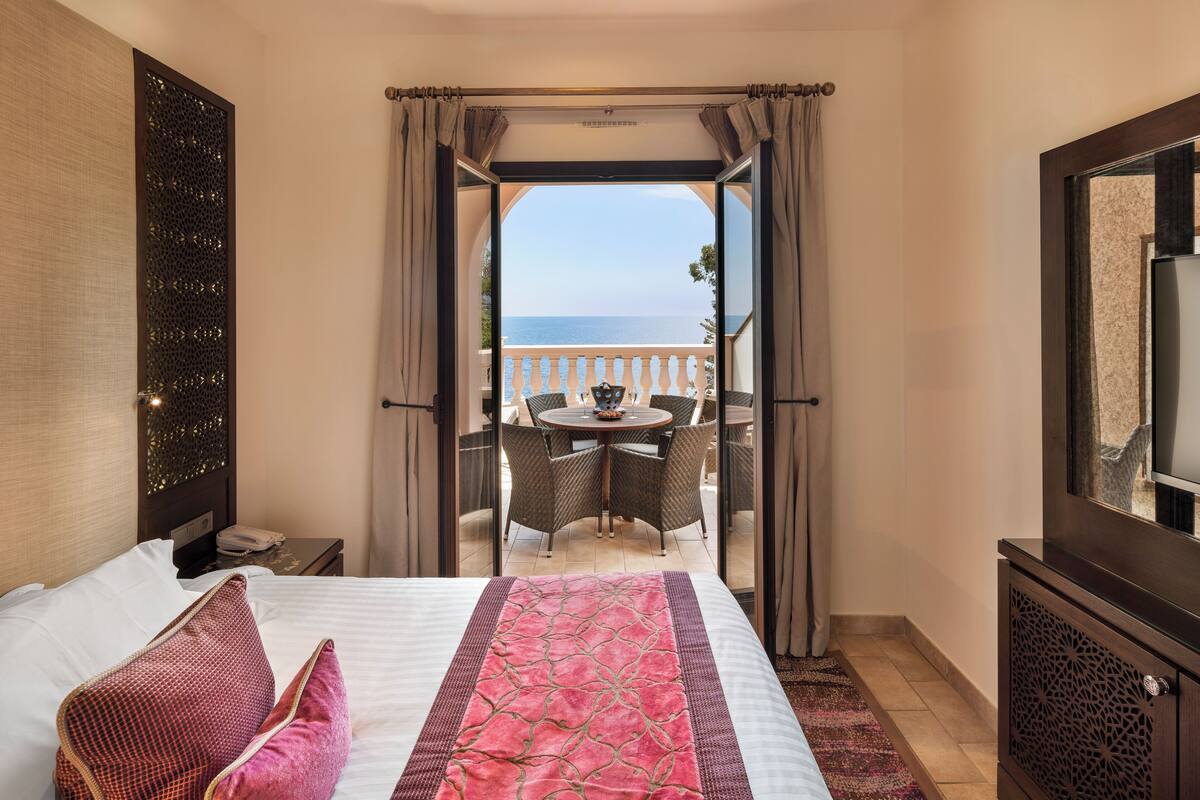 A hotel room with a bed, a view of the sea through an open door, a balcony with chairs, and a TV on the wall.