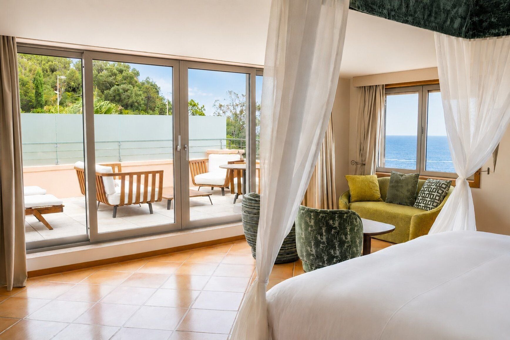 A bedroom with a large bed, a balcony with outdoor furniture, and a view of the sea.