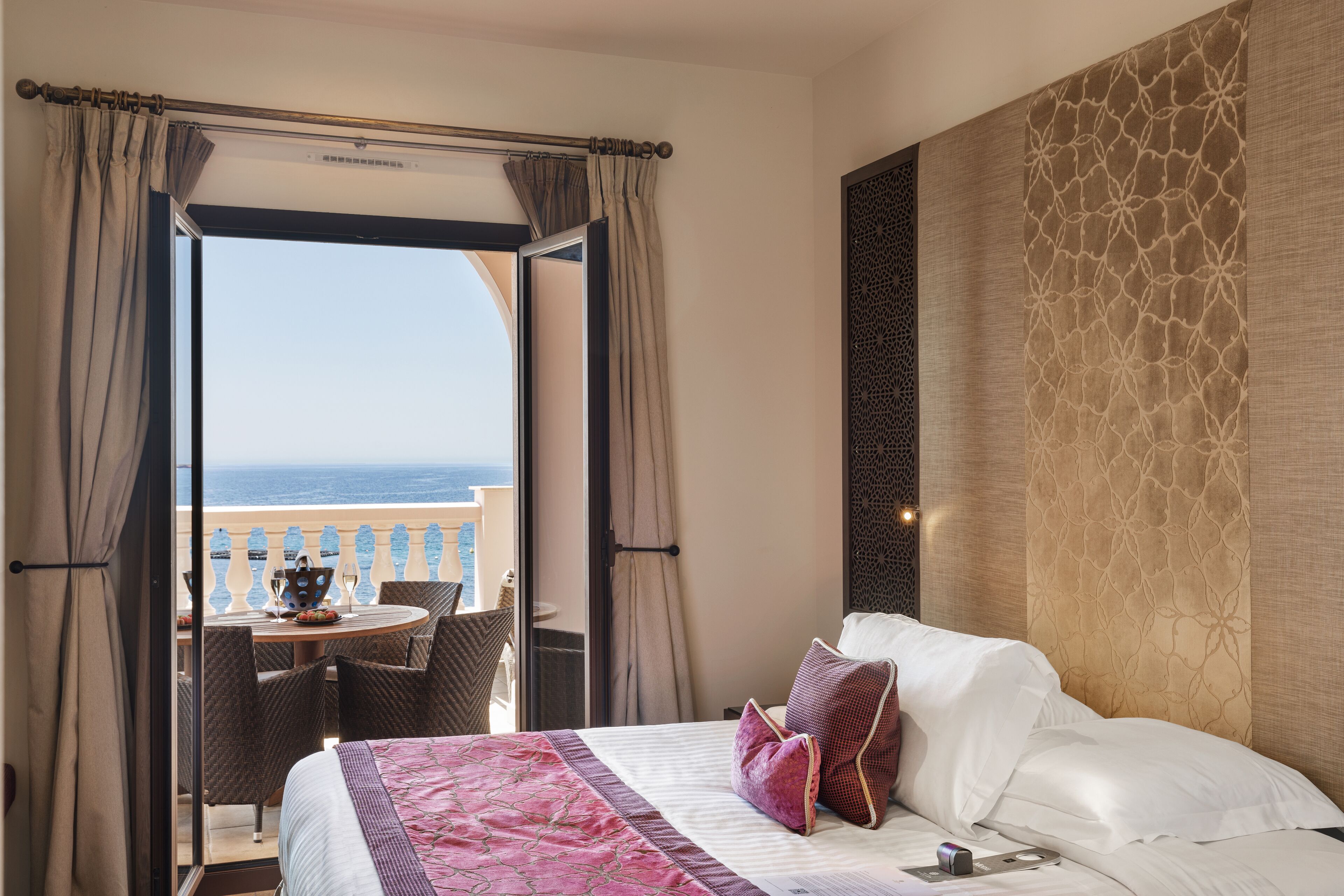 Een hotelkamer met balkon, een bed met witte lakens, een deken met roze en witte patronen en uitzicht op zee.