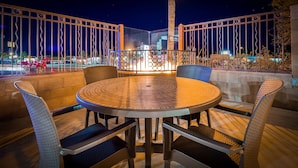 An outdoor seating area with a round table and chairs, a metal fence, and a view of the city at night.