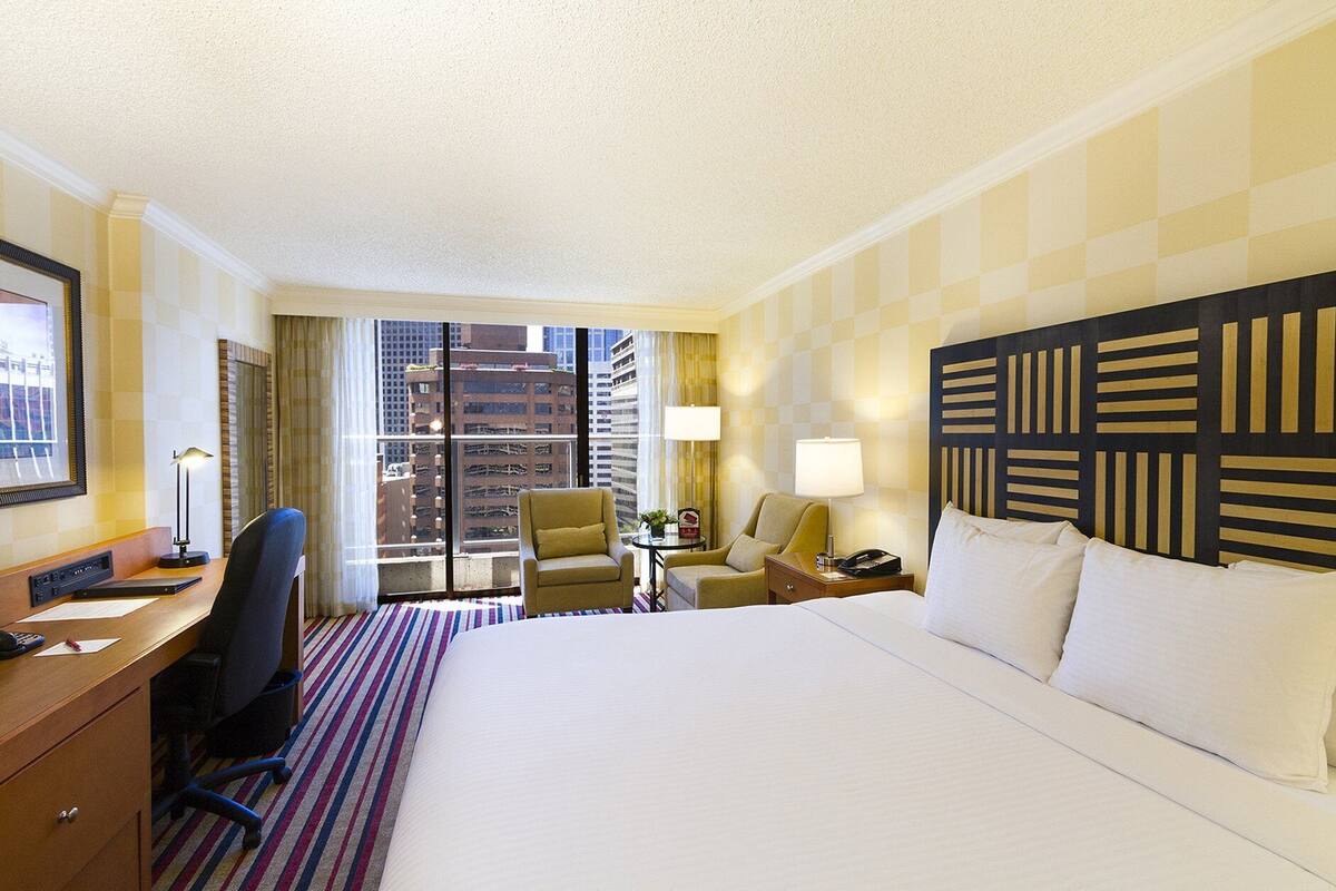 A hotel room with a large bed, a desk with a computer, a chair, a lamp, and a view of the city through the window.