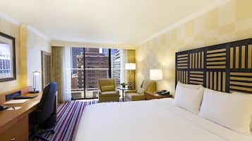 A hotel room with a large bed, a desk with a computer, a chair, a lamp, and a view of the city through the window.