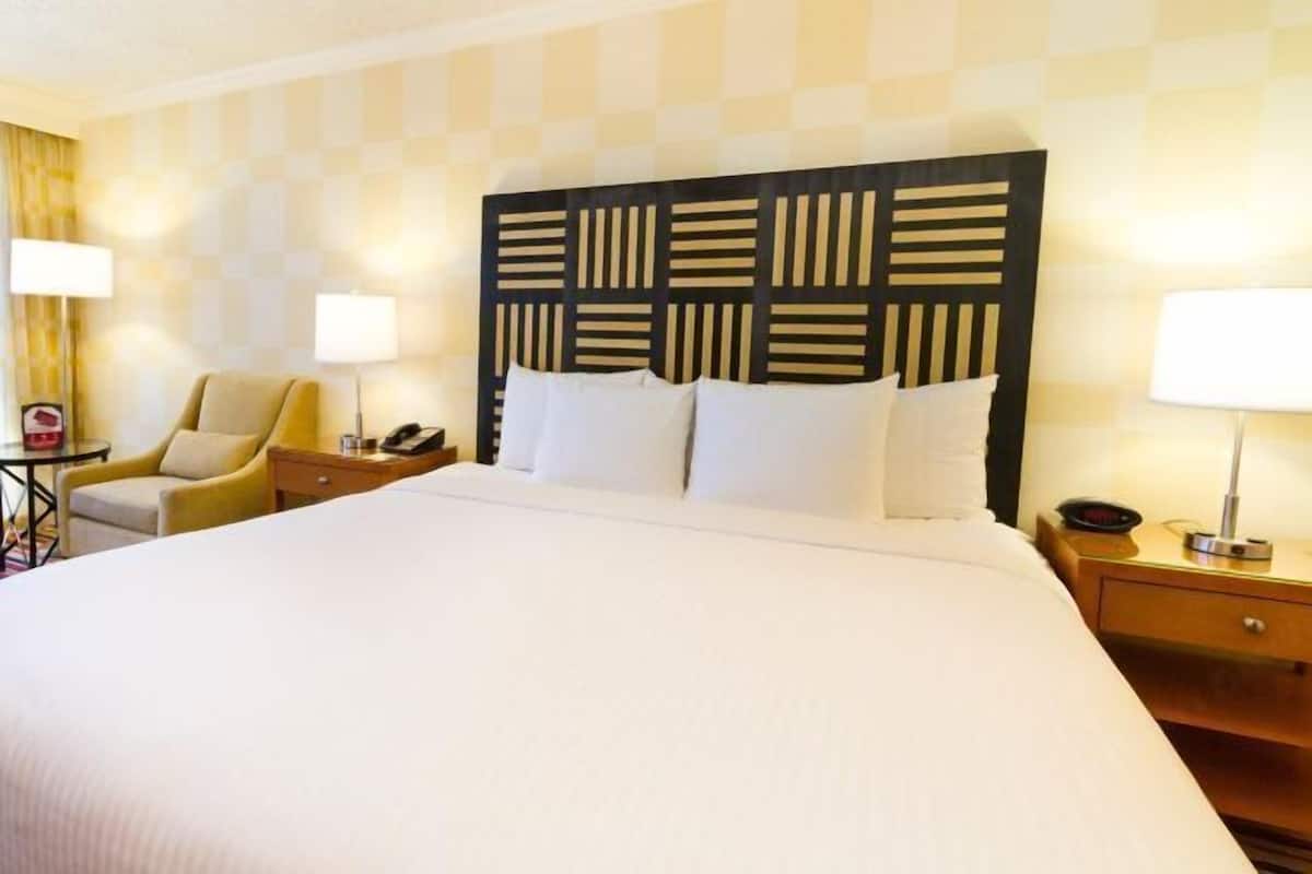 A hotel room with a large bed, a headboard with a geometric pattern, two bedside lamps, a chair, and a nightstand with a phone.