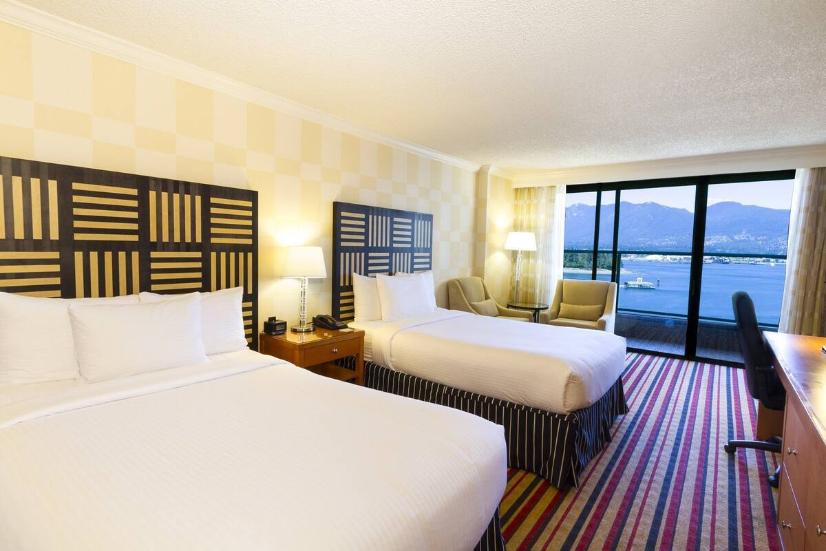 A hotel room with two beds, a desk, a chair, and a view of a body of water and mountains.