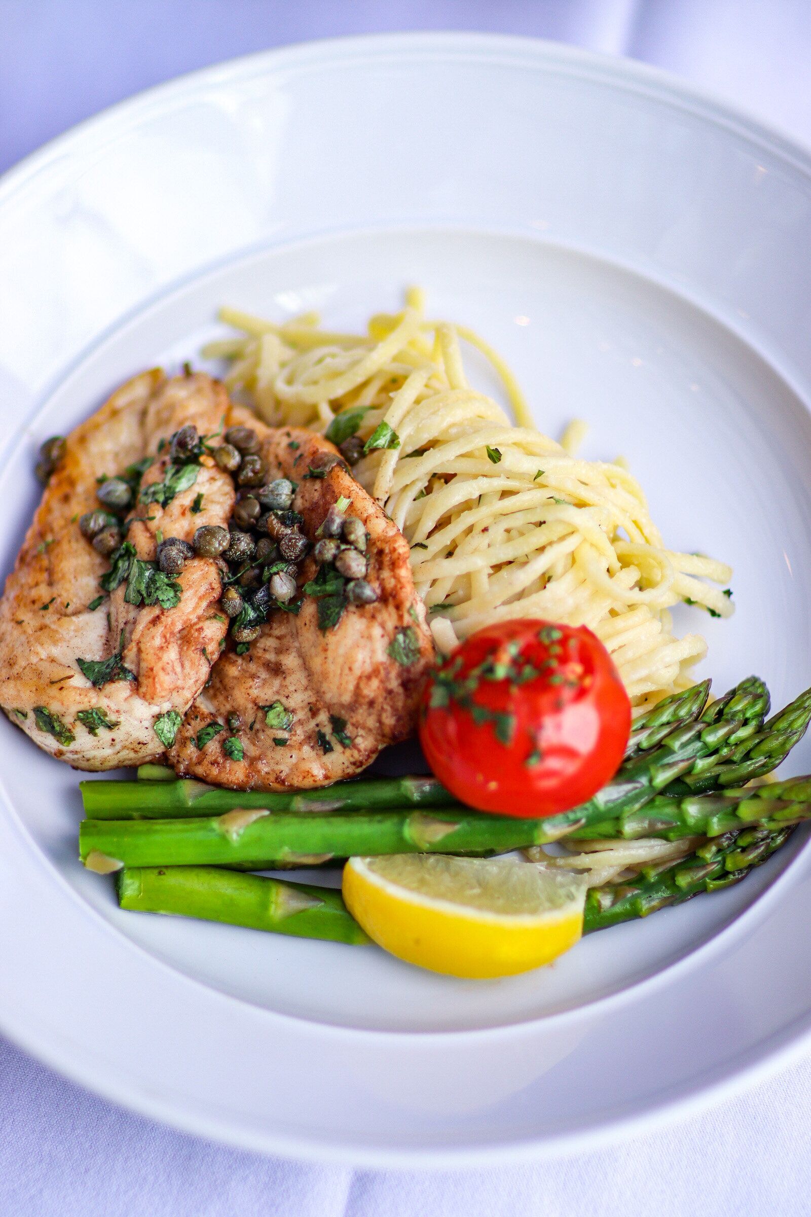 A plate with grilled chicken, spaghetti, asparagus, a cherry tomato, and capers.