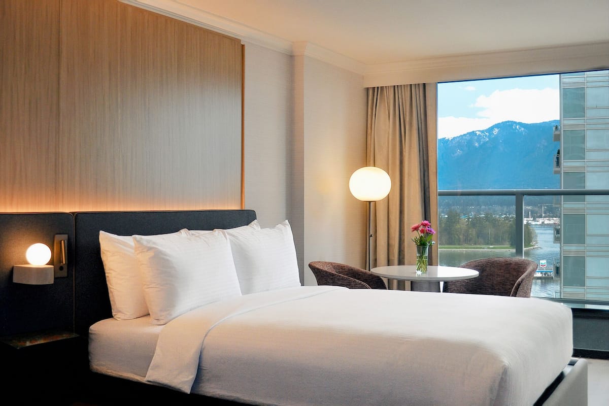 A hotel room with a bed, a nightstand with a lamp, a small table with a vase, and a view of mountains through a window.