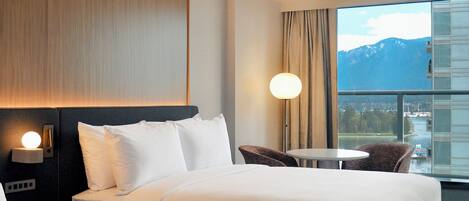 A hotel room with a bed, a nightstand with a lamp, a chair, and a large window with a view of mountains.