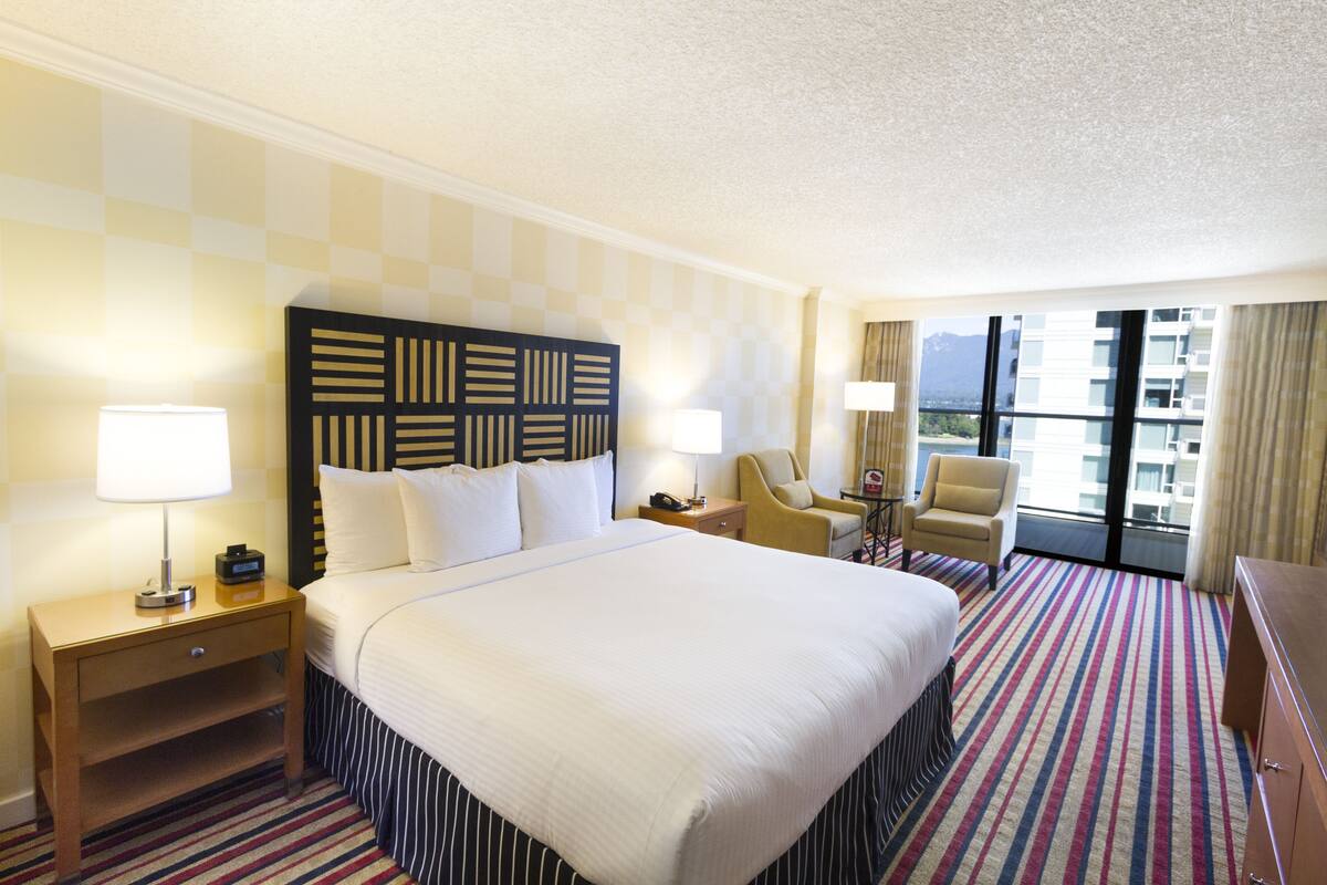 A hotel room with a large bed, two bedside tables, a desk, a chair, and a view of the city through large windows.
