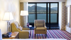 A hotel room with a large window offering a view of a city and mountains, a bed, two armchairs, a small table, and a lamp. - Pinnacle Hotel Harbourfront (Vancouver)