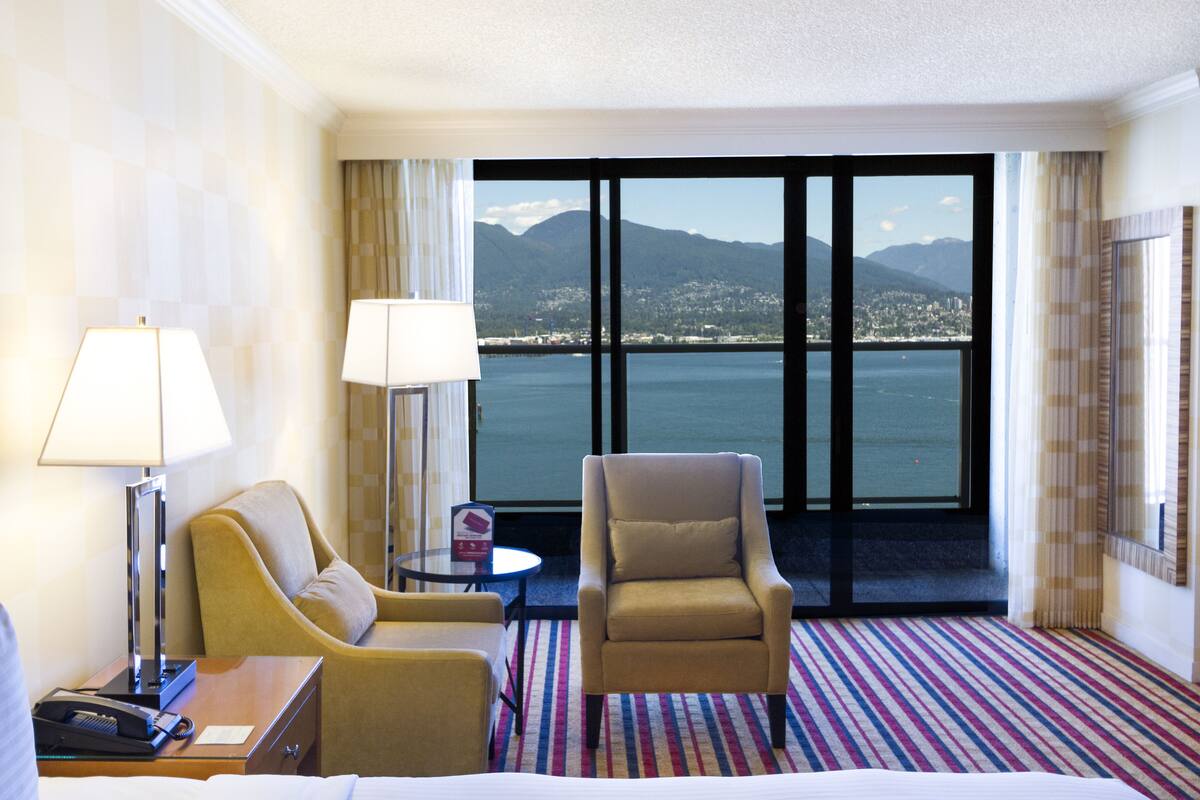 A hotel room with a large window offering a view of a city and mountains, a bed, two armchairs, a small table, and a lamp.