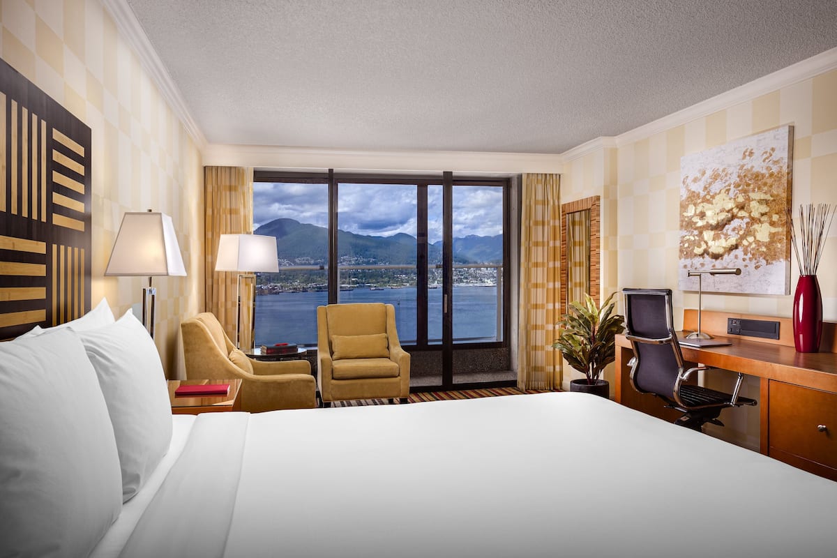 A hotel room with a large bed, a desk with a chair, a lamp, and a view of mountains and water.