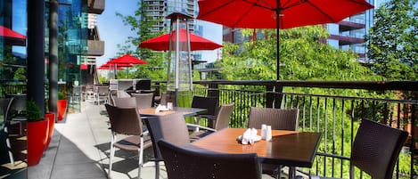 An outdoor patio with tables and chairs, red umbrellas, and a view of a cityscape.