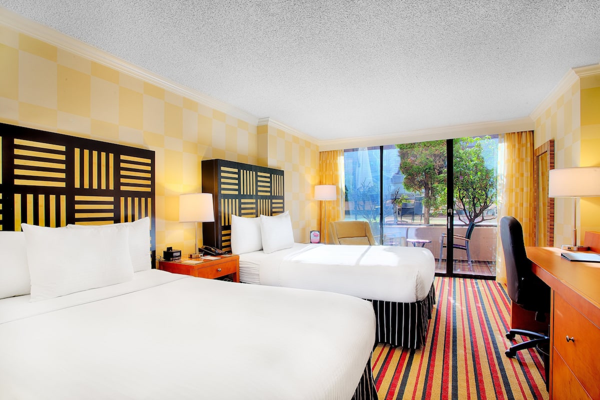 A hotel room with two beds, a desk, a chair, and a view of the outdoors.
