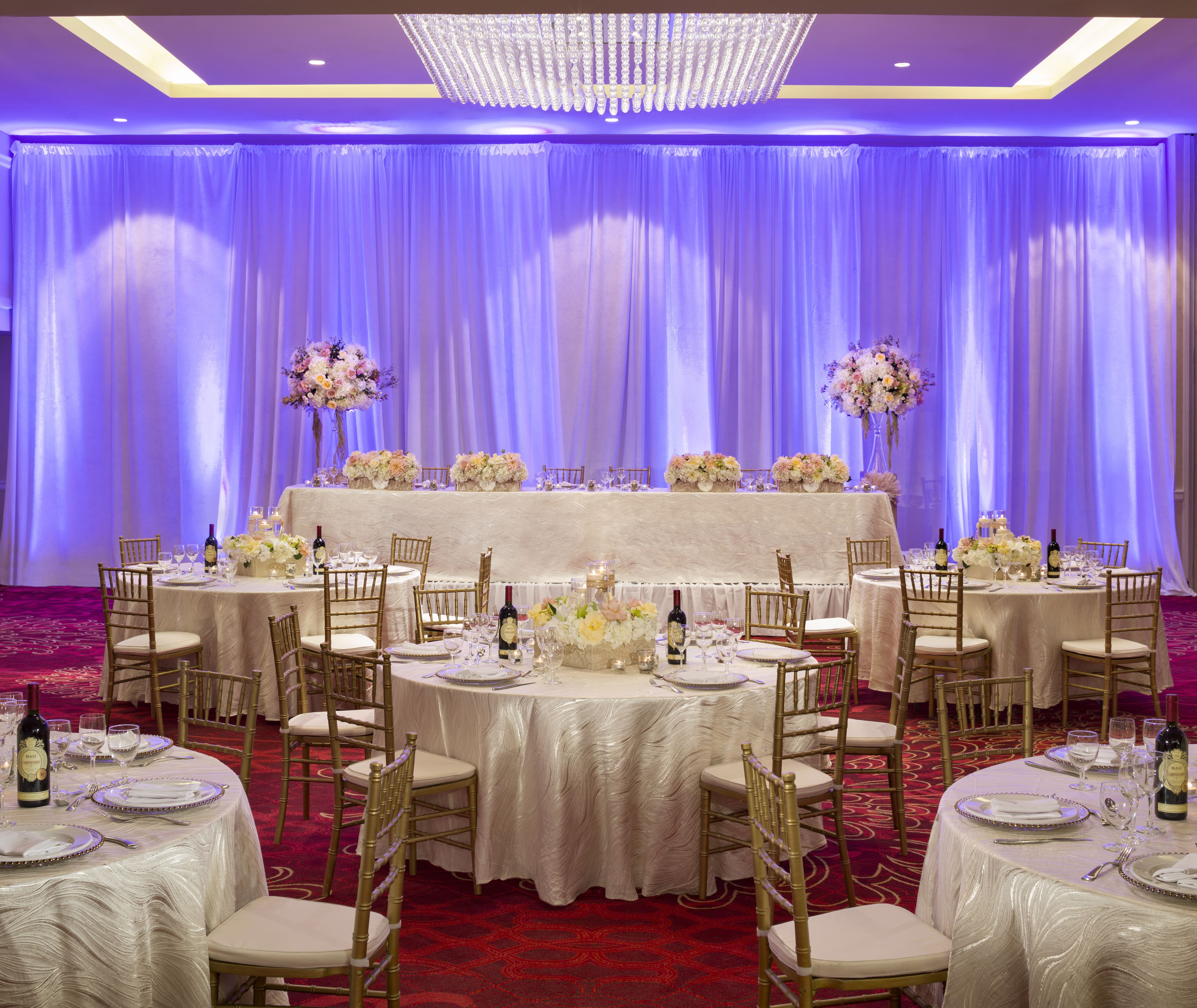 A banquet hall with round tables set for a formal event, featuring white tablecloths, gold chairs, and floral centerpieces.