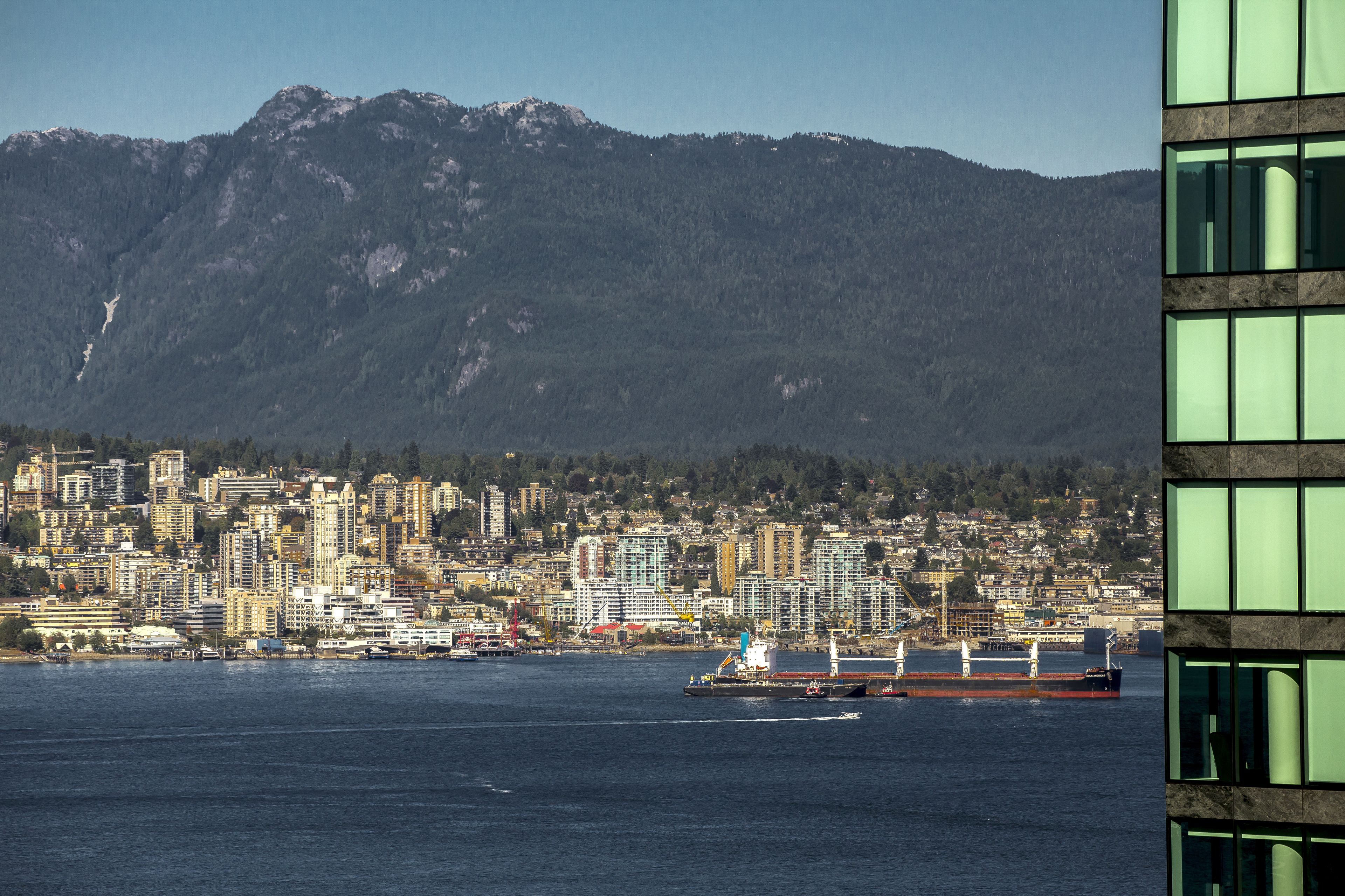 Un paisaje urbano frente al agua, con un barco grande atracado en el puerto, una montaña al fondo y un edificio moderno con ventanas reflectantes.