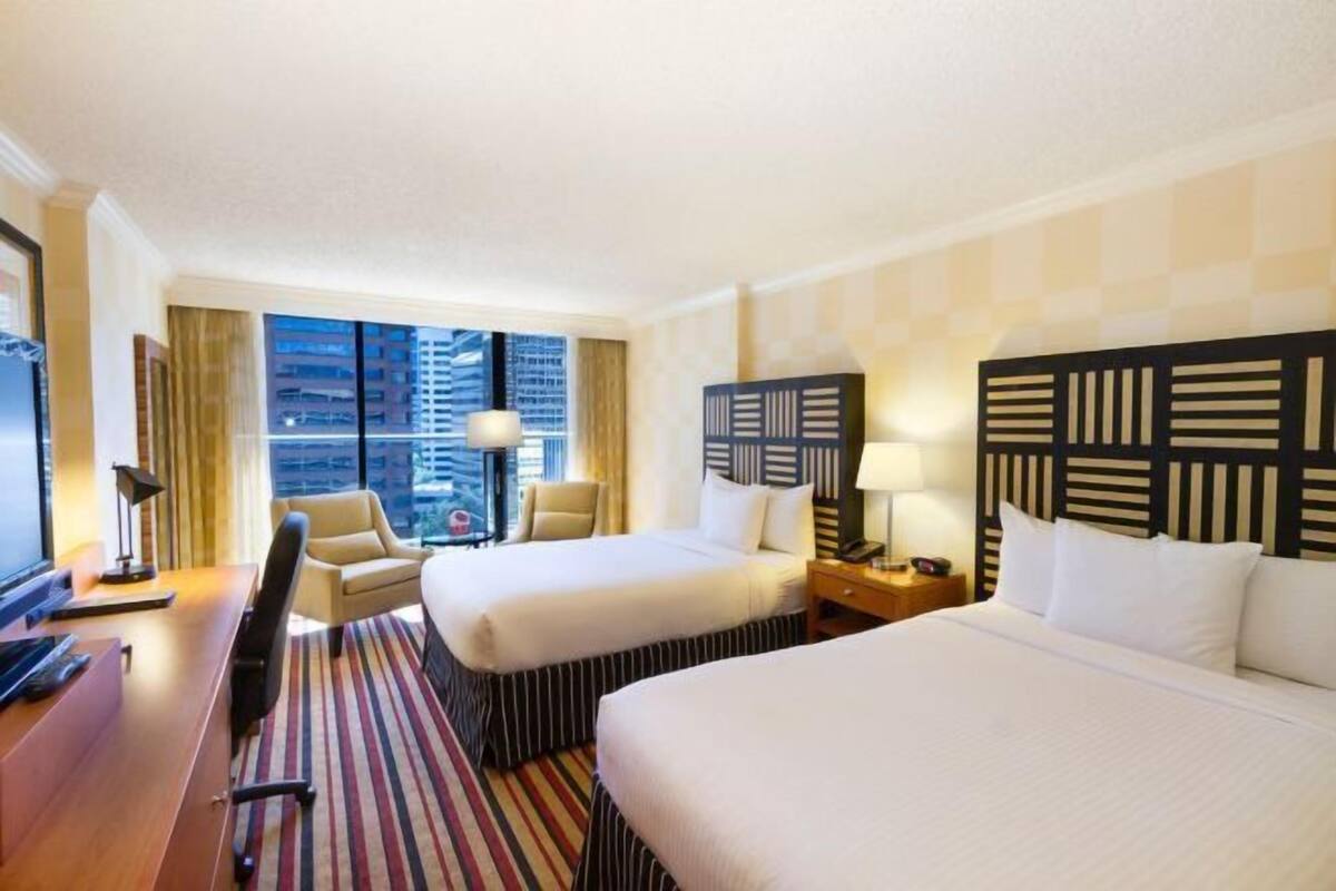 A hotel room with two beds, a desk, a chair, a TV, and a view of the cityscape.