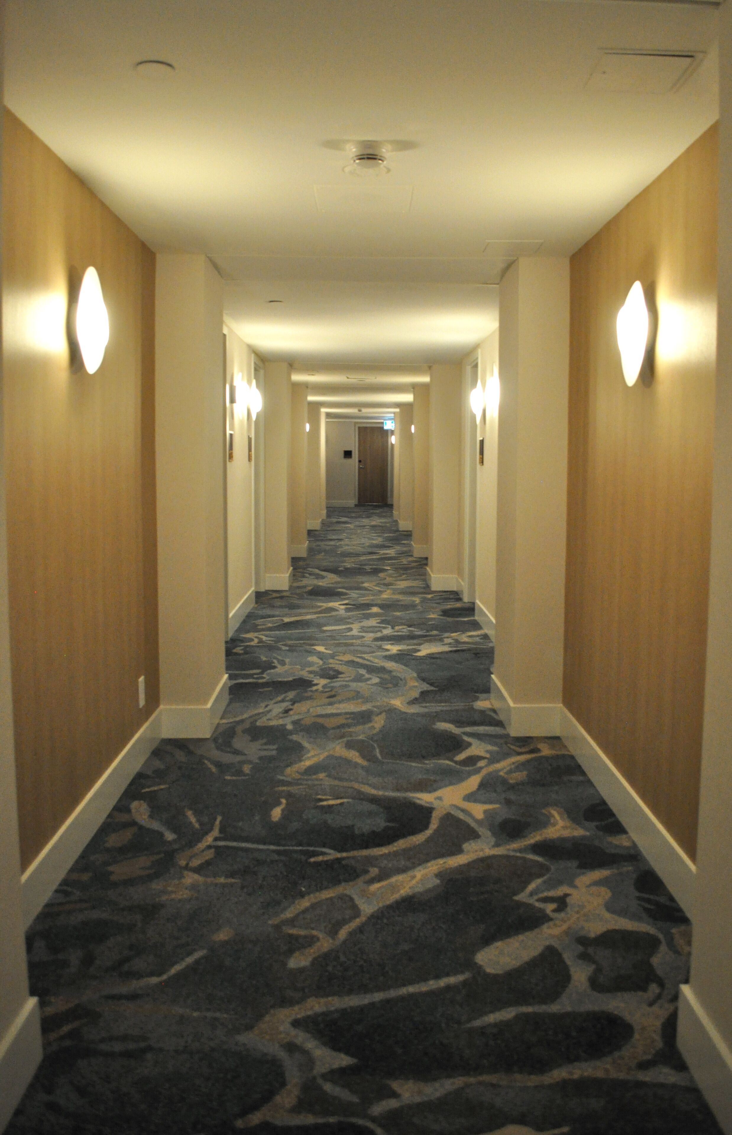A hotel hallway with a patterned carpet, wooden walls, and wall sconces.