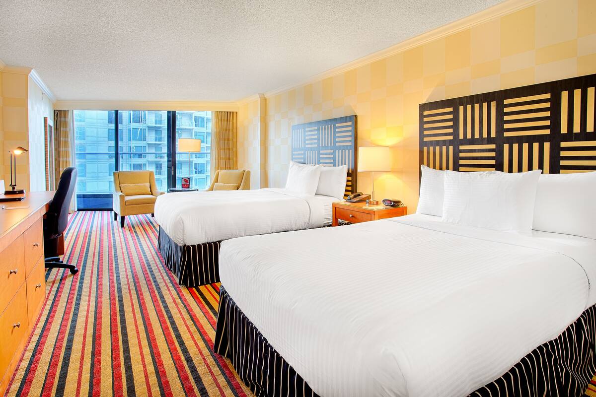 A hotel room with two beds, a desk, a chair, and a view of the city.