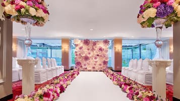A hotel corridor with a white carpet leading to a reception area, flanked by white chairs and tables, and adorned with large floral arrangements.