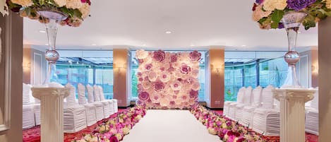 A hotel corridor with a white carpet leading to a reception area, flanked by white chairs and tables, and adorned with large floral arrangements.