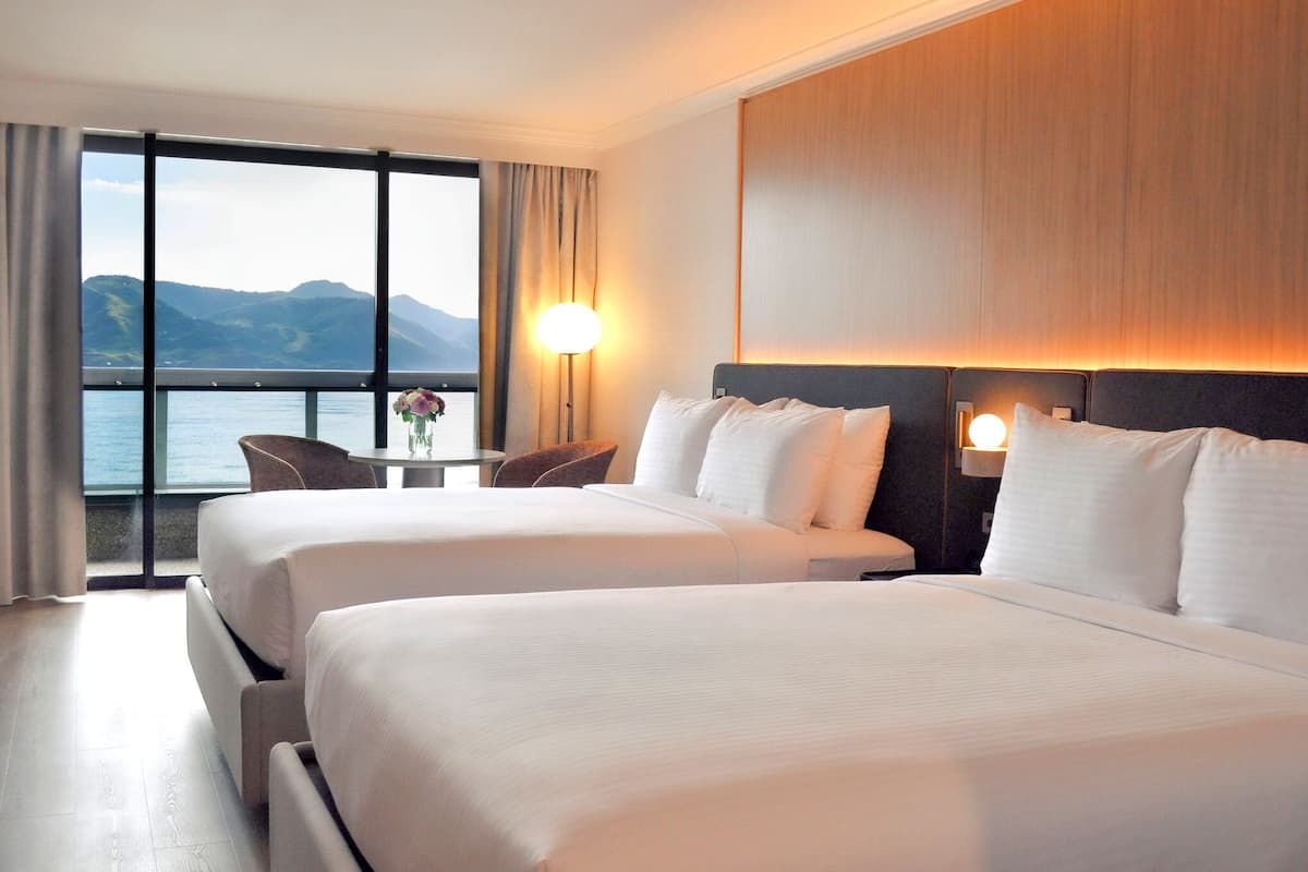 A hotel room with two beds, a view of the sea, a table with a vase, and a lamp.